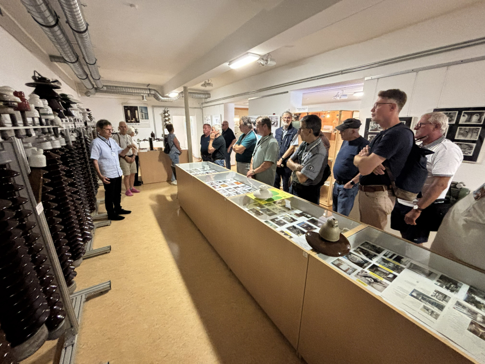 Eine Gruppe von Menschen steht in einem Ausstellungsraum und hört einer Person zu, die etwas erklärt. In Vitrinen und Regalen sind technische Objekte zu sehen.