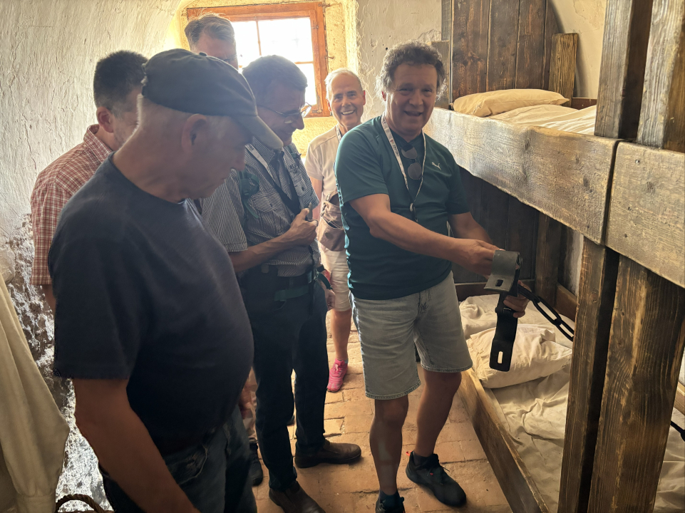 Mehrere Menschen stehen in einem historischen Raum vor einem hölzernen Etagenbett und schauen sich die Einrichtung gemeinsam an.