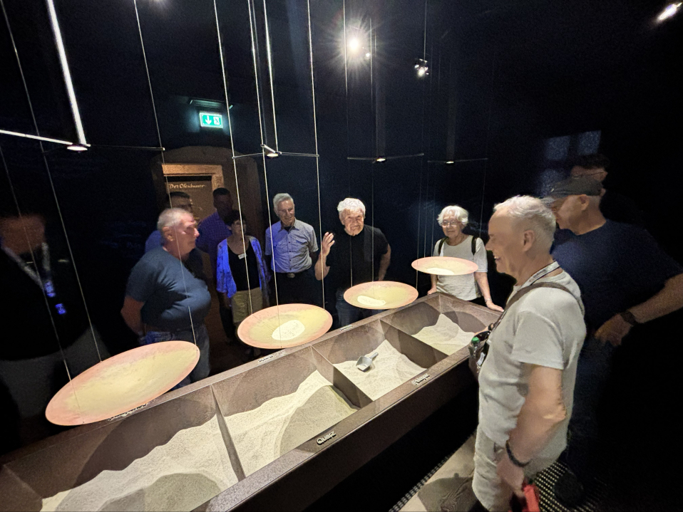 Eine Gruppe von Menschen steht in einem dunklen Ausstellungsraum um ein beleuchtetes Exponat und betrachtet die Präsentation gemeinsam.