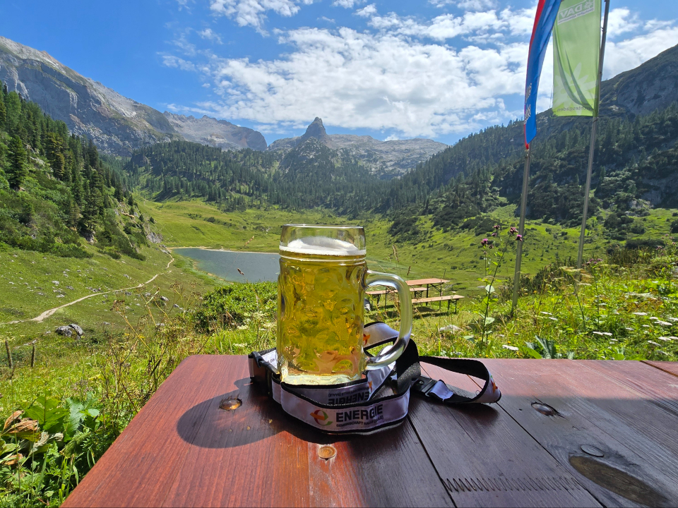 Ein Bierkrug steht auf einem Holztisch vor einer Berglandschaft mit See, Wiesen und bewaldeten Hängen unter blauem Himmel.
